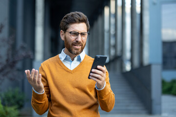 A man in glasses, looking at his phone with a confused expression, gestures with his hands in an...