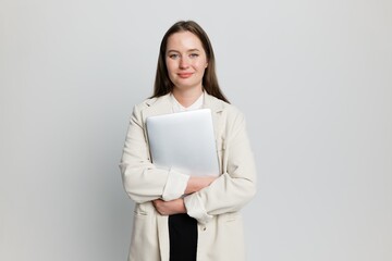 Confident young woman in a blazer holding a laptop while standing against a neutral backdrop in a...