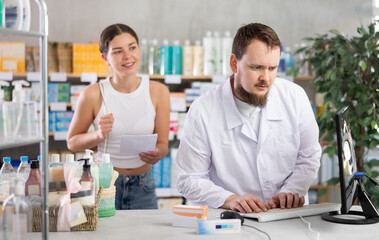 Obraz premium Young man pharmacist looking for prescription on computer to young woman buyer in pharmacy