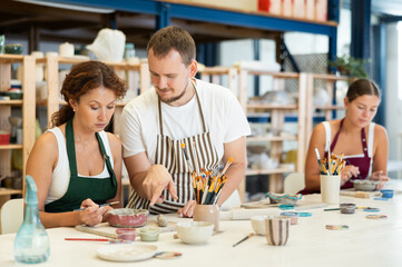 Student sits near desktop and paints clay product in presence of man teacher. Lover of applied art...