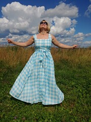 Woman in a blue and white dress is standing in a field. She is wearing sunglasses and has her arms...