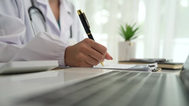A professional in a white coat writes on a clipboard while using a laptop, suggesting healthcare administration, medical documentation, or online consultation.