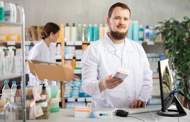 Adult male pharmacist holds a package of pills in hands and stands behind the counter near the...