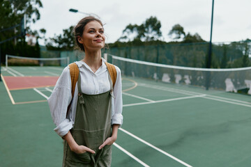 Obraz premium Young woman in casual overalls and a backpack, smiling confidently on a tennis court during a cloudy day, representing an active and healthy lifestyle