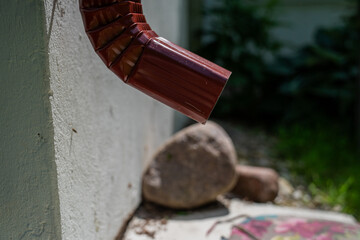 red metal rain gutter downspout attached to white stucco wall with rocks below in backyard garden...
