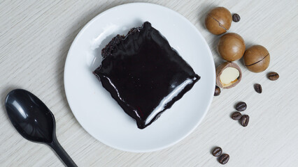 Chocolate cake featuring glossy icing resting on white plate, scattered coffee beans and macadamia...