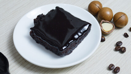 Chocolate brownie with glossy chocolate icing sits on a white plate next to macadamia nuts and coffee beans, creating a tempting dessert presentation