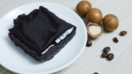Piece of freshly baked chocolate brownie, covered with melted chocolate, is served on a white plate next to macadamia nuts and coffee beans, creating a tempting dessert presentation