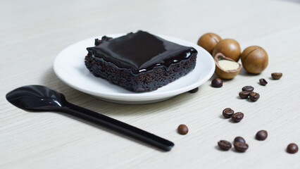 Rich chocolate brownie sitting on white plate, accompanied by scattered coffee beans, macadamia nuts across rustic wooden surface, presenting decadent dessert composition
