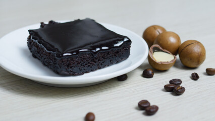 Square chocolate brownie with a glossy chocolate topping sits on a white plate, accompanied by macadamia nuts and coffee beans on a light wooden surface, creating a tempting dessert scene