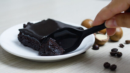 Hand uses a black plastic spoon to take a bite of a rich, dark chocolate brownie on a white plate,...