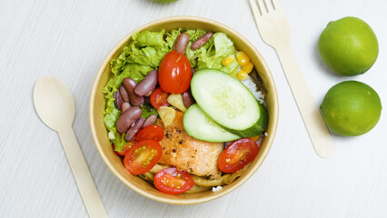 Grilled salmon poke bowl with rice, lettuce, cucumber, cherry tomatoes, beans, corn and two limes resting on a white table next to wooden cutlery, promoting a healthy and balanced diet