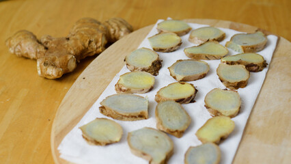 Thinly sliced ginger root is drying on a white paper towel placed on a wooden cutting board, with a whole ginger root visible in the background on the wooden table