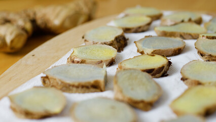 Thinly sliced ginger root drying on a white paper towel, resting atop a wooden cutting board, offers a flavorful ingredient for cooking or a natural remedy for wellness