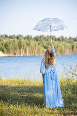 Woman with an umbrella in the summer heat. Woman in a blue dress standing by the lake. Blue water, beautiful nature and view