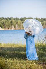 Woman with an umbrella in the summer heat. Woman in a blue dress standing by the lake. Blue water, beautiful nature and view
