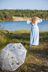 Woman with an umbrella in the summer heat. Woman in a blue dress standing by the lake. Blue water,...