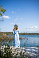 Woman standing on the edge of a lake. Woman in a blue dress and with a wreath on her head. Blue water, beautiful nature and view. Water nymph. Mavka