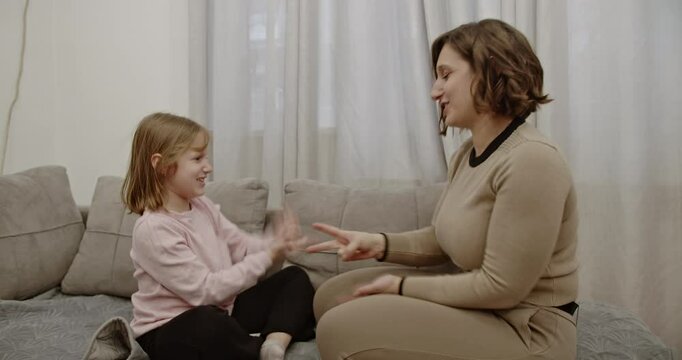 A mother and daughter are sitting on a couch in their living room, playing a hand-clapping game and smiling.