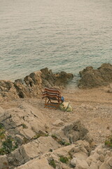 A wooden folding chair draped with a striped towel and a blue cloth stands on a rocky beach facing the calm sea.