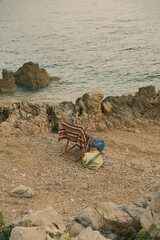A wooden folding chair draped with a striped towel and a blue cloth stands on a rocky beach facing the calm sea.