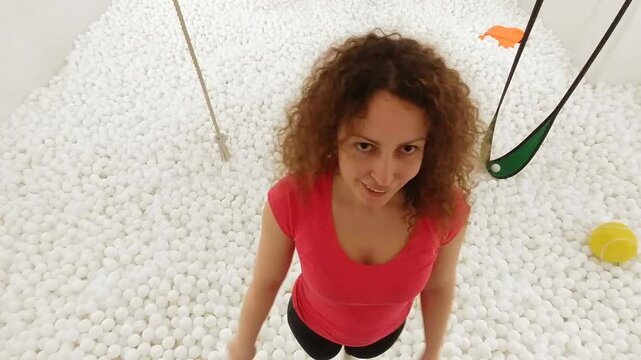 Woman is swinging in a ball pit. She is wearing a pink shirt. The ball pit is filled with white balls