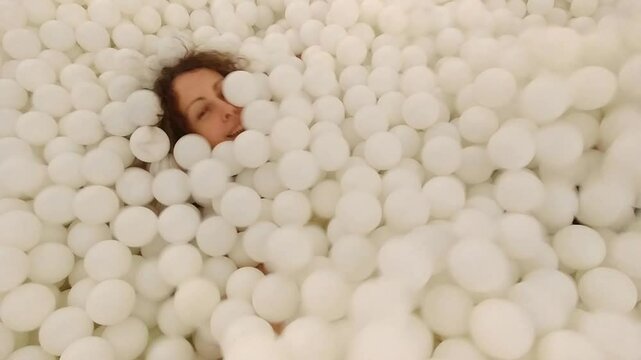 Woman is in a white ball pit. She is covered in white balls. The balls are scattered all over her body