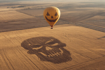 A skull drawn in the fields of an open field, seen from above with a hot air balloon painted like a Halloween decoration, with a scary face flying over it. 