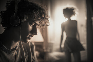 A curly-haired man wearing headphones is listening to music in the bedroom, and the silhouette of an attractive woman can be seen dancing behind him. 