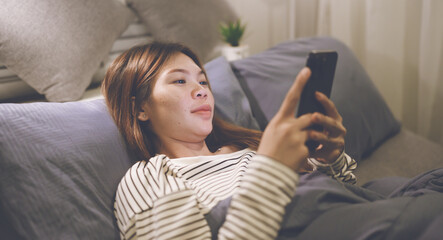 A young woman lies awake at night in bed, scrolling on her glowing phone, battling insomnia,...