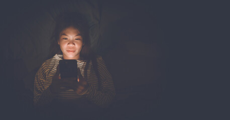 A young woman lies awake at night in bed, scrolling on her glowing phone, battling insomnia,...