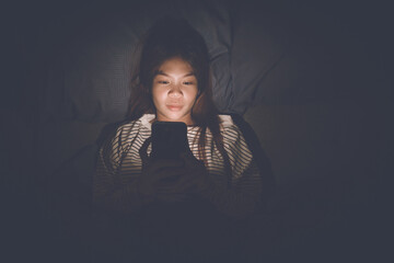 A young woman lies awake at night in bed, scrolling on her glowing phone, battling insomnia,...