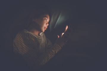 A young woman lies awake at night in bed, scrolling on her glowing phone, battling insomnia,...