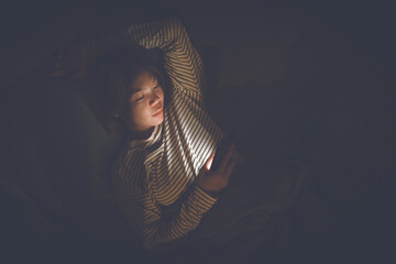 A young woman lies awake at night in bed, scrolling on her glowing phone, battling insomnia,...