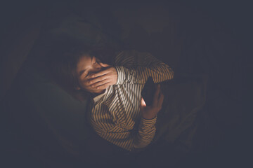 A young woman lies awake at night in bed, scrolling on her glowing phone, battling insomnia,...