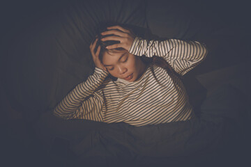A young woman lies in bed, covering her ears with her hands, looking distressed, suggesting she's...