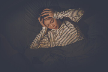 A young woman lies in bed, covering her ears with her hands, looking distressed, suggesting she's...