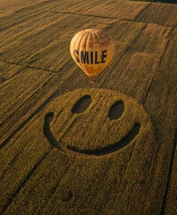 A hot air balloon with the word "SMILE" written on it flies over an endless field, leaving behind shadows in the shape of a smiley face. 