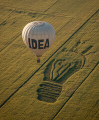 A hot air balloon with the word "IDEA" on it floats over an open field, where there is a large drawing of a light bulb's shadow on the green grass.