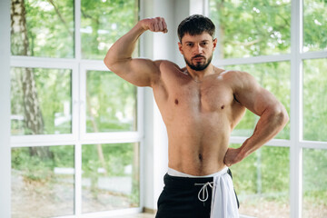 Shows the muscles. Shirtless strong man is standing against panoramic windows
