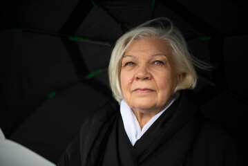 Elderly woman under black umbrella looking ahead with serious expression.