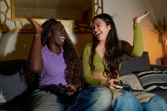 Two women sitting on a cozy sofa, joyfully playing video games together and sharing laughter in a relaxed evening atmosphere
