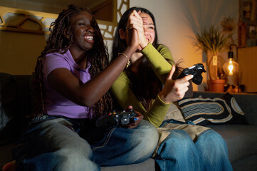 Two women are sitting on a couch, playing videogames with gamepads and giving each other a high...
