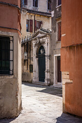Sunlit Venetian Alley with Ornate Classical Doorway. Narrow stone street framed by weathered buildings and an ornate, classical doorway.