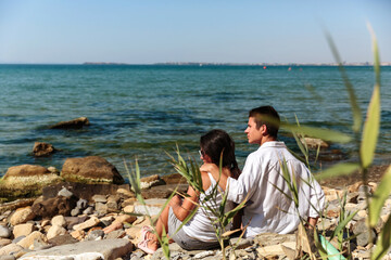 A teenage boy and his mother sit on a rocky beach surrounded by green plants, gazing at the vast...