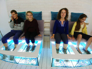four people are sitting on chairs next to each other. fish in aquariums do peeling legs pedicure....