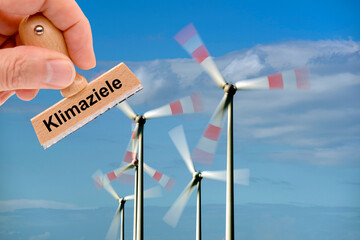Ein Holzstempel ist beschriftet mit dem Aufdruck Klimaziele und wird von einer Hand vor eine Windkraftanlage mit drehenden Rotorblättern gehalten - FOTOMONTAGE