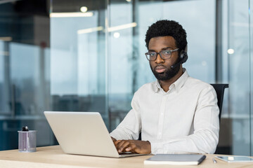 Fototapeta premium An African-American man wearing a headset and glasses works on a laptop at a desk in a modern office setting.