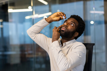 A man applies eye drops while looking up in a modern office setting, highlighting healthcare and...