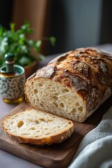 Freshly baked rustic loaf of bread with a single slice cut on a wooden cutting board, with a blurred background including an oil bottle and green plant, evoking a cozy, homely feeling